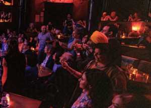 People in a dimly lit club or theater are watching a performance by Toby Lightman, some standing and clapping while others sit at tables with drinks and candles.